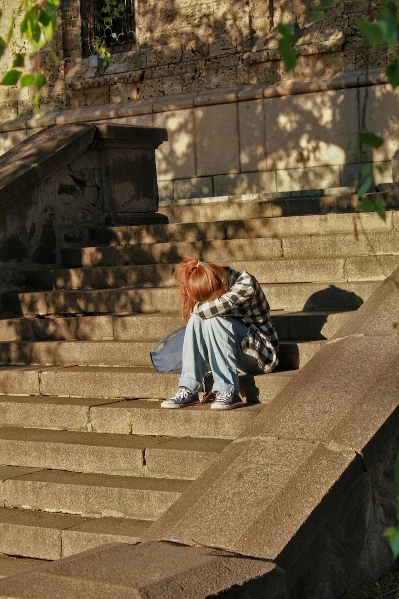 Jeune personne assise, tête dans les bras sur des escaliers en pierre Une jeune personne, seule et découragée, symbolisant le refus scolaire et la phobie scolaire.